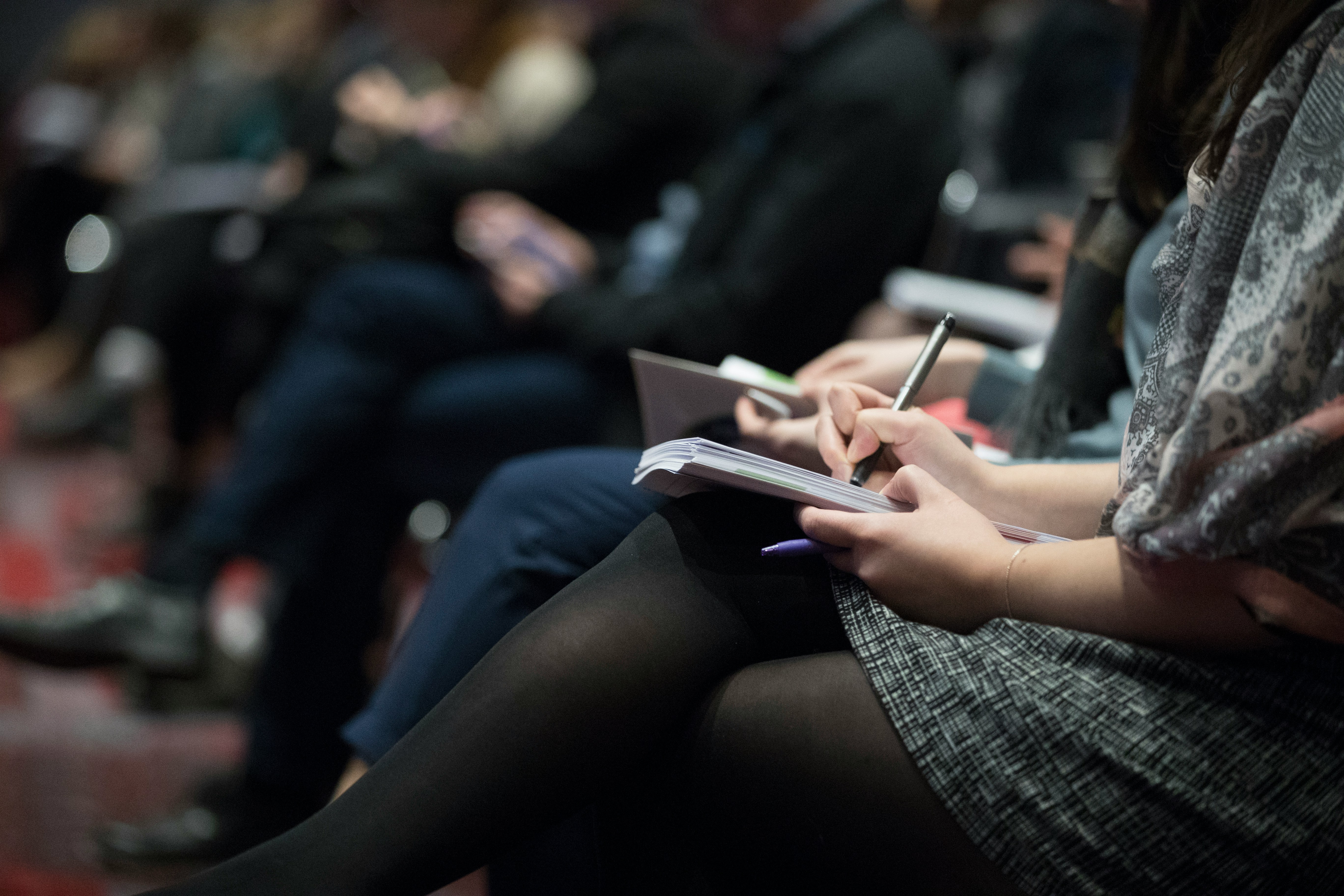 A woman writes on a notepad on her lap. Other people can be seen out of focus in the background similarly writing.